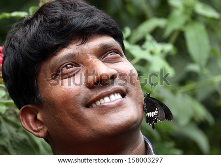 Handsome young Indian smiling with butterfly on face - concept of nature conservation. The photos shows a happy adult indian male with a just born black butterfly sitting on his face