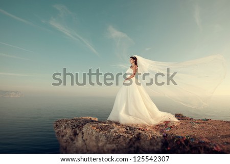 beautiful bride stands on a cliff above the sea in a glamorous white wedding dress