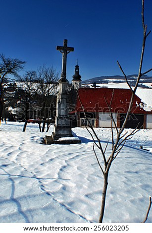 crucifix with house and church on the background in Metylovice village during nice winter day