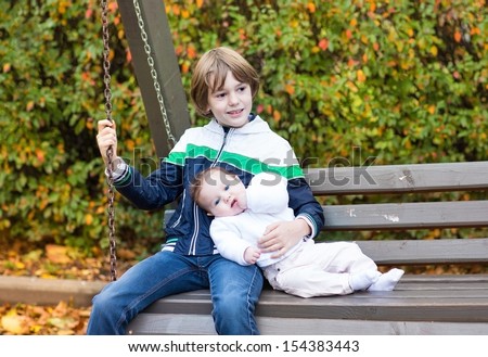Little boy and his newborn baby sister relaxing on a wooden swing in a colorful garden with red, yellow and golden trees