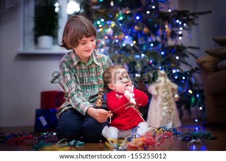 Beautiful little baby girl and her cute brother playing together under a Christmas tree in a dark living room