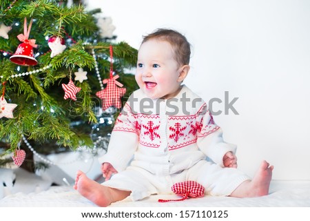 Funny laughing baby girl playing under a red and white decorated Christmas tree wearing a knitted jacket