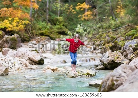 Smiling happy boy trying to cross a wild mountain river jumping on stones in a beautiful autumn forest
