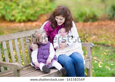 Young mother relaxing on a wooden bench in a colorful autumn park with her toddler daughter and newborn son covered with a white knitted warm blanket