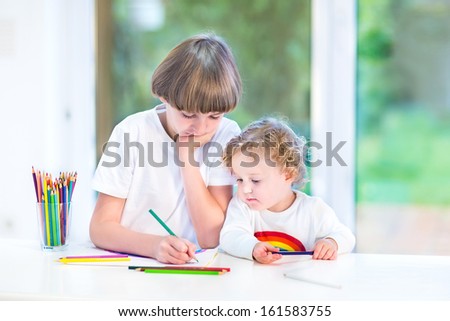 Brother and little toddler sister having fun together painting at a white desk next to a big window with garden view