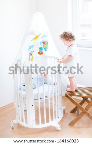 Cute toddler girl standing on a chair and watching her newborn baby brother sleeping in a white round crib with colorful toys and canopy