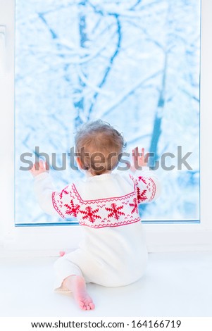 Little baby sitting at a window watching snow covered trees in the winter garden