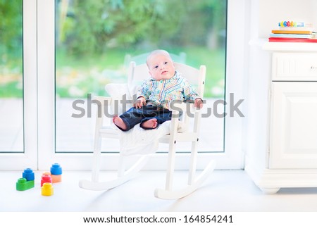 Funny newborn baby boy in a white rocking chair next to a big window with garden view