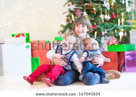 Three happy children - cute boy, his toddler sister and a newborn baby - sitting under a beautiful Christmas tree