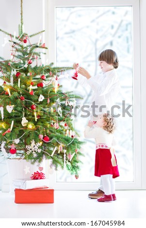 Adorable toddler girl with curly hair wearing a warm red dress helping her brother to decorate a beautiful Christmas tree standing next to a big window with a view of a snowy garden