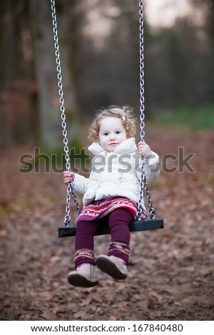 Adorable toddler girl with curly hair wearing a white warm jacket and purple dress having fun on a swing in a dark autumn park on a cold November day