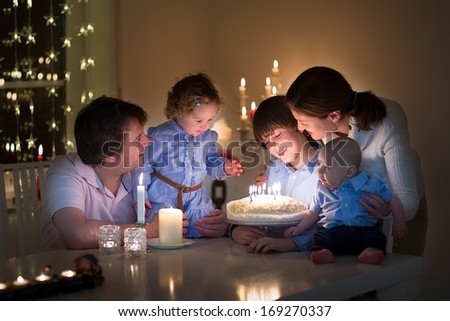 Young happy family with three kids celebration the birthday of their son blowing out the candles on a cake in a dark living room