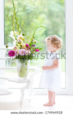 Beautiful toddler girl watching flowers in a big vase next to a window with garden view