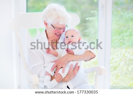 Loving grandmother singing a song to her newborn baby grandson sitting in a white rocking chair next to a big garden view window