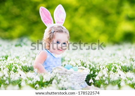 Adorable toddler girl wearing bunny ears playing with Easter eggs in a white basket sitting in a sunny garden with first white spring flowers