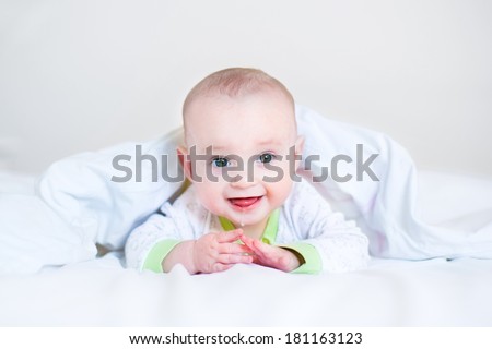 Adorable funny laughing baby playing peek-a-boo under a white blanket in a sunny bedroom waking up in the morning