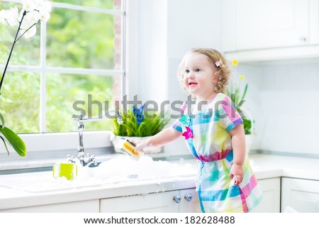 Cute curly toddler girl in a colorful dress washing dishes, cleaning with a sponge and playing with foam in the sink in a beautiful sunny white kitchen with a garden view window in a modern home