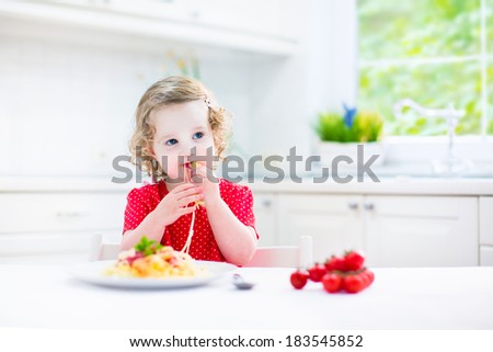 Cute curly laughing toddler girl in a red shirt playing with fork and spoon eating spaghetti with tomato sauce and vegetables for healthy lunch sitting in a white sunny modern kitchen with big window
