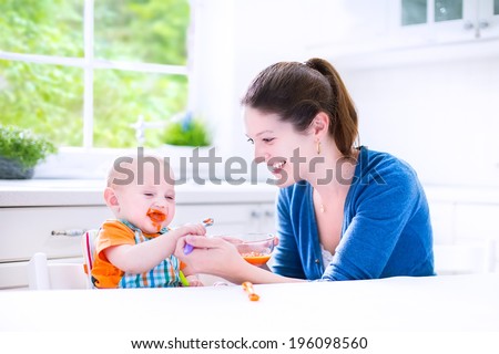 Young attractive mother feeding her cute baby son, giving him his first solid food, healthy vegetable pure from carrot with a plastic spoon sitting in a white sunny kitchen at a window at home