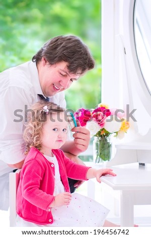 Young happy father in a business suit and tie brushing the hair of his daughter, cute little curly toddler girl, sitting on a white dresser with a beautiful round mirror in a bedroom with a window