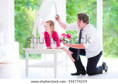 Young happy father in a business suit and tie brushing the hair of his daughter, cute little curly toddler girl, sitting on a white dresser with a beautiful round mirror in a bedroom with a big window