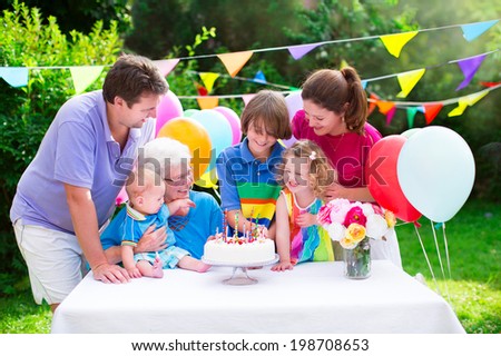 Happy big family - young parents, grandmother, three kids, teenage boy, toddler girl and little baby celebrating birthday party with cake and candles in the garden decorated with balloons and banners