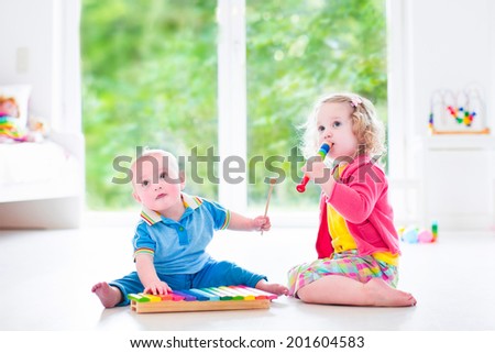 Two little children - cute curly toddler girl and a funny baby boy, brother and sister playing music, having fun with colorful xylophone and flute at a window; kids early development class