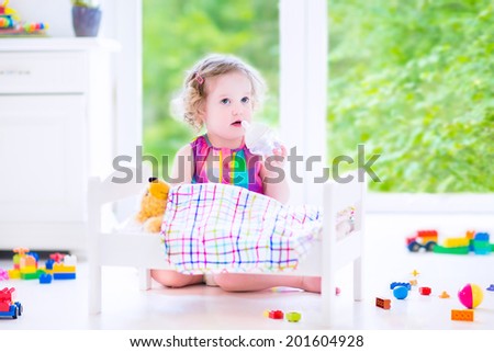 Cute curly toddler girl in a colorful dress feeding her toy bear in a white crib playing in a sunny bedroom with big garden view windows