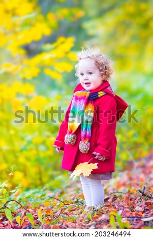 Funny little toddler girl in a red jacket and colorful knitted hat and scarf playing with golden maple leaves in a sunny park with yellow and orange trees on a warm autumn day