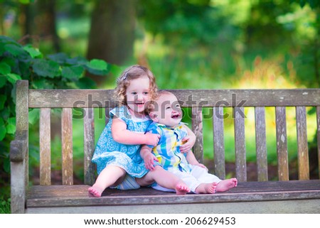 Adorable kids, little curly girl and a cute baby boy, brother and sister, sitting together on a wooden bench in a garden, hugging and kissing, relaxing and having fun on a sunny summer day