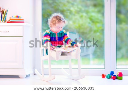 Cute curly little girl, funny toddler wearing a warm colorful knitted dress reading a book relaxing in a white rocking chair next to a big garden view window at home or daycare center
