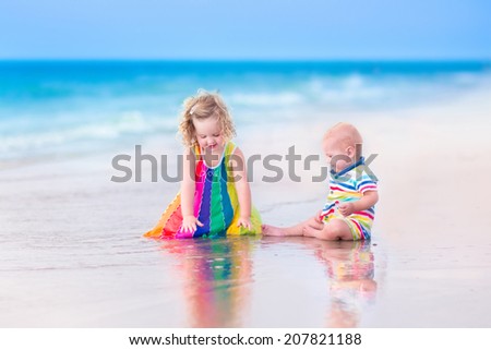Two happy children, cute curly toddler girl and funny baby boy, brother and sister, playing together digging sand in water on a beautiful beach enjoying a hot summer vacation day on a tropical island