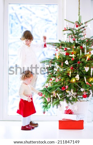 Happy children playing next to a Christmas tree. Beautiful curly toddler girl in a warm knitted dress helping her brother to decorate the Xmas tree standing next to a big window to a snowy garden