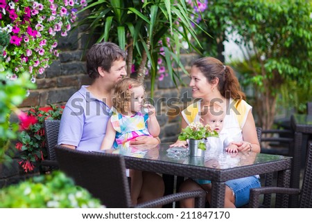 Happy young family, parents with two children, adorable little girl and a funny baby boy, eating lunch in a beautiful outdoor cafe with flowers in a city center on a warm summer day