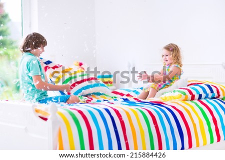Two children, happy laughing boy and cute curly little girl having fun at pillow fight with feathers in the air jumping, laughing and giggling in a white bedroom with colorful bedding