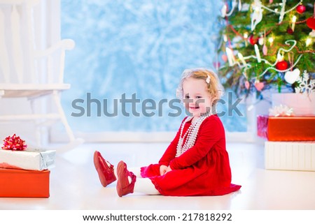 Cute curly little girl in a red dress and white pearl necklace playing under Christmas tree with presents sitting on the living room floor with rocking chair next to a window into snowy winter garden