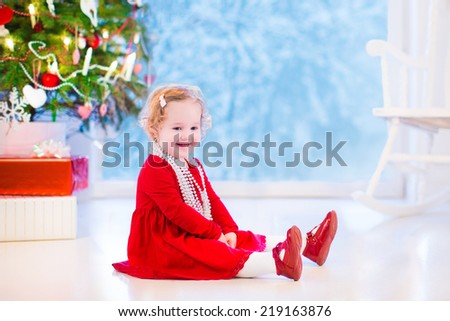 Cute curly little girl in a red dress and white pearl necklace playing under Christmas tree with presents on the floor of a white living room with rocking chair next to window into snowy winter garden