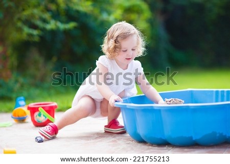Cute curly toddler girl in a white summer dress playing in a sand box enjoying a warm sunny day in the garden