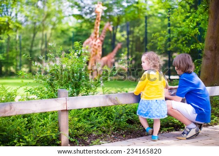 Happy school boy and his toddler sister cute little girl with curly hair wearing a dress having fun together in a zoo watching giraffes and other animals on a day trip during summer vacation