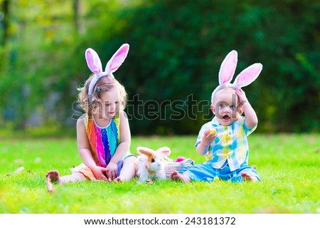 Two little children, cute curly toddler girl and funny baby boy wearing bunny ears having fun at Easter egg hunt playing with basket and toy rabbit in a sunny spring garden
