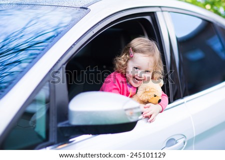Cute laughing curly toddler girl playing with toy teddy bear sitting in a silver color modern family car on front seat watching out a window in a side mirror enjoying weekend vacation ride after rain