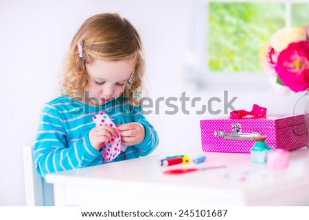 Cute creative little girl sewing a dress for her teddy bear doll, playing with needles and ribbons in a white sunny room at home or preschool