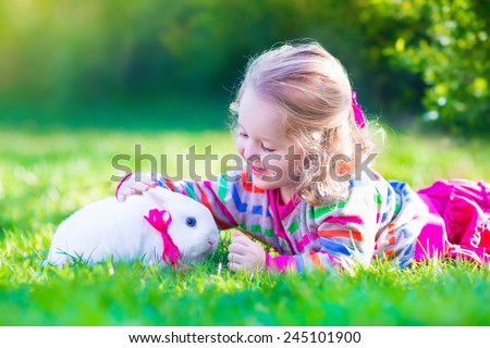 Adorable curly toddler girl playing with a real rabbit in a sunny summer garden, child feeding bunny a carrot.