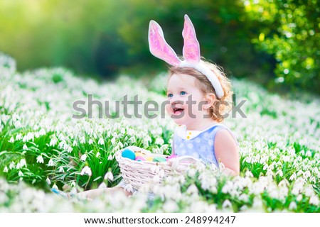 Adorable curly toddler girl wearing bunny ears playing with Easter eggs in a white basket sitting in a sunny garden with first white spring flowers