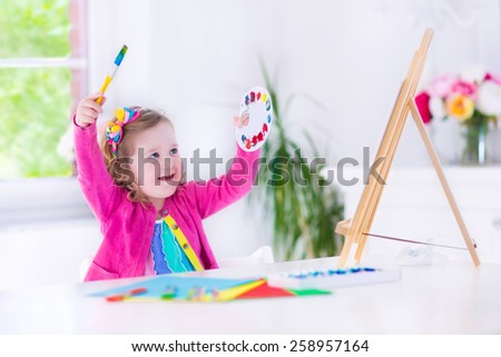 Cute happy little girl, preschooler, painting with water color on canvas standing on easel in a sunny white room at home or elementary school, creative young artist at work. Kids paint. Children draw.