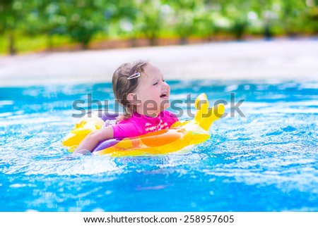 Adorable little girl wearing a colorful sun protection swimming suit playing with water splashes at beautiful pool in a tropical resort having fun during family summer vacation