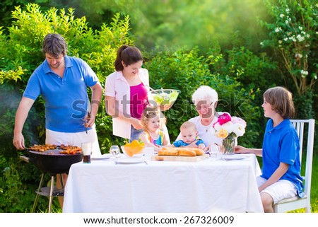 Grill barbecue backyard party. Happy big family, mother, father, teen son, cute toddler daughter and baby, enjoying BBQ lunch with grandmother eating grilled meat in the garden with salad and bread.