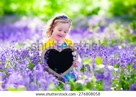 Child playing in bluebells forest. Little girl holding a wooden heart shape chalk board standing in a park with beautiful spring bluebell flowers. Copy space for your text. Kids having fun outdoors.