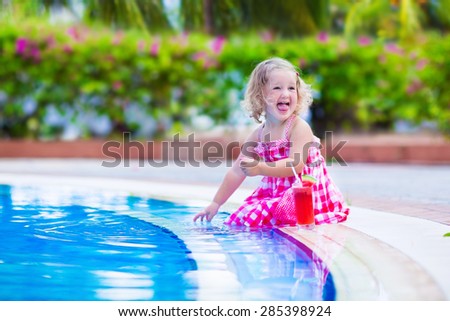 Beautiful little girl, cute toddler with curly hair wearing red summer dress, sitting at a swimming pool drinking water melon juice with fresh fruit having fun at family vacation in a tropical resort