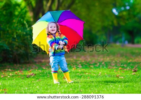 Child with colorful umbrella playing in the rain. Kids play outdoors by rainy weather. Toddler kid in coat and waterproof boots jumping in autumn garden. Fall fun for children, raining in the park.